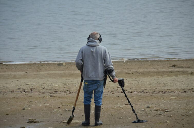 A man walks on a beach with a metal detector and shovel.